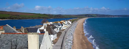 The image is a landscape photograph of Slapton Sands beach on the coast of Devon, England, in the United Kingdom. The scene shows the long, sweeping stretch of Slapton Sands beach from an elevated viewpoint, likely taken during the late morning in early spring. To the left, traditional houses with slate rooftops line the edge of the village, while a narrow road runs parallel to the shoreline, separating the dwellings from the sand. The beach extends into the distance, bordered by the vivid blue sea on one side and the freshwater lake of Slapton Ley on the other, with rolling green hills visible further inland. The clear sky suggests a bright spring day, adding clarity to the coastal features and the rural landscape of this region in Devon, England.
